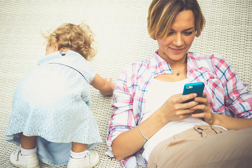 Woman lays on back using smart phone while infant crawls next to her