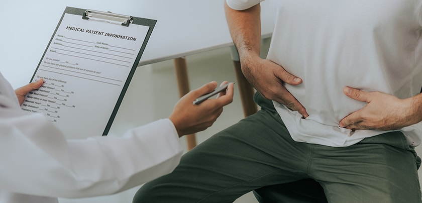 Stock image of man pointing to his stomach with dr seated across looking at a clipboard. 