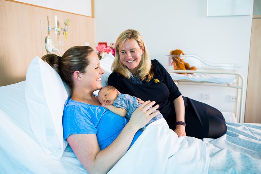 A midwife perches next to a new mother holding her newborn in a hospital bed.