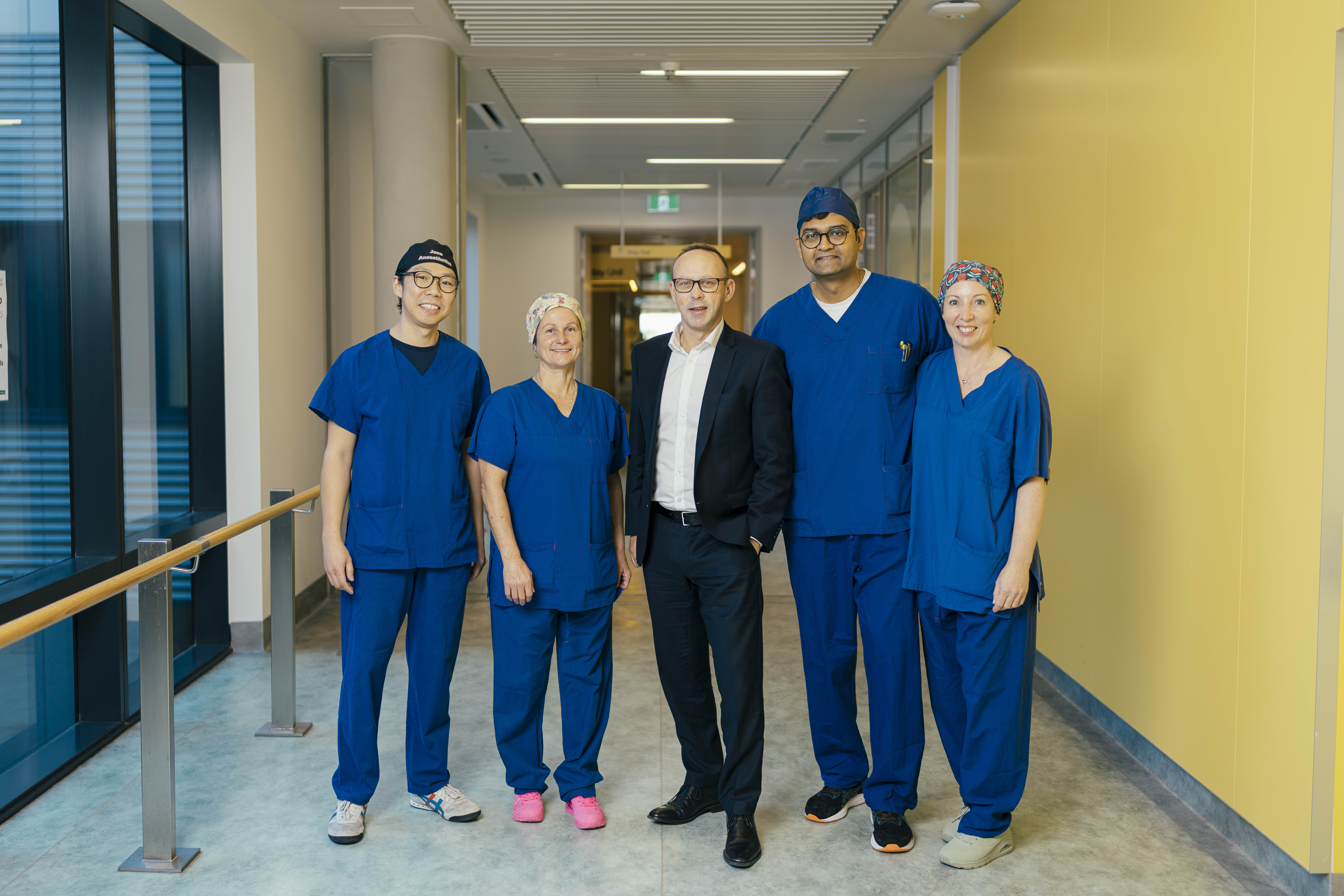 A man in a suite is standing in a hospital corridor, flanked by 2 medical professionals on each side