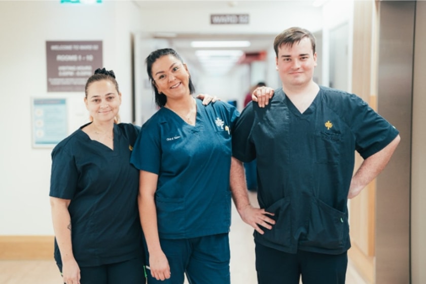 Three nurses in blue scrubs stand in a hospital corridor, posing and smiling in front of the camera