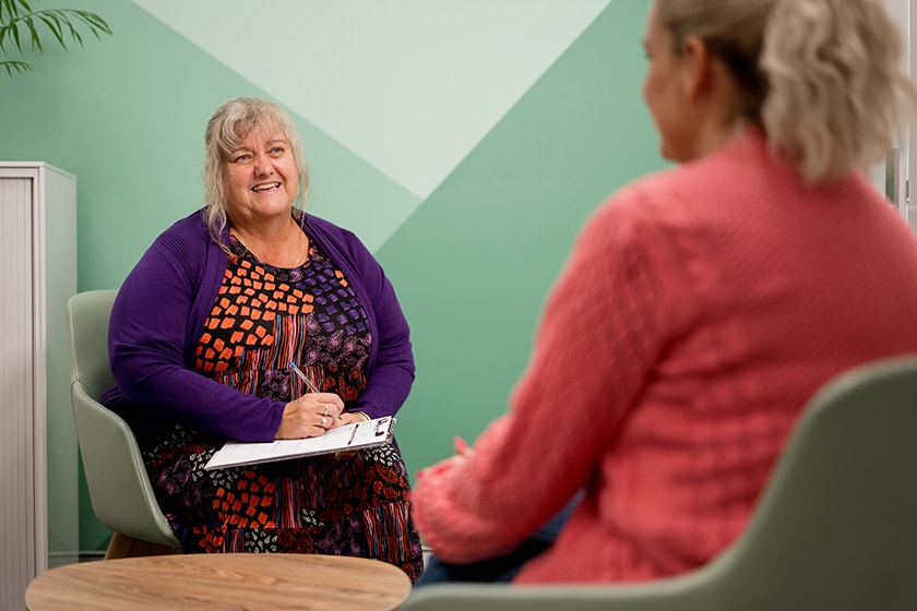 a woman in a purple dress sits on a chair before a light green wall, talking to a woman in a pink sweater who has her back to the camera