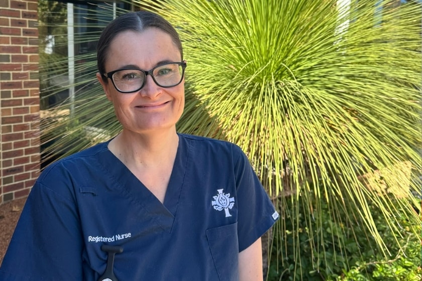 A woman with classes in a blue scrub with the words registered nurse on, is standing outside in front of a green bush, smiling at the camera