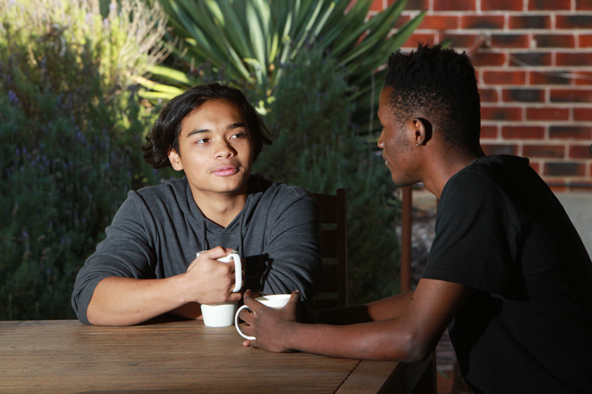 Two youth persons sitting in an outdoor alfresco setting facing one another whilst sitting at a wooden table with coffee mugs in their hands.
