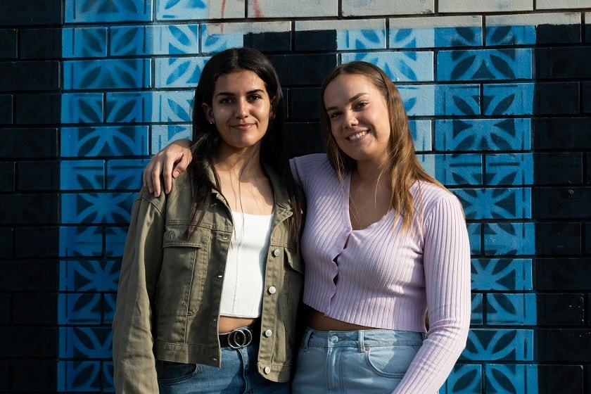 Two young women with their arms around each other, smiling at camera.
