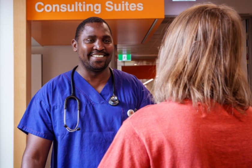 A smiling healthcare professional in blue scrubs with a stethoscope talks with a patient in a hospital corridor near consulting suites.