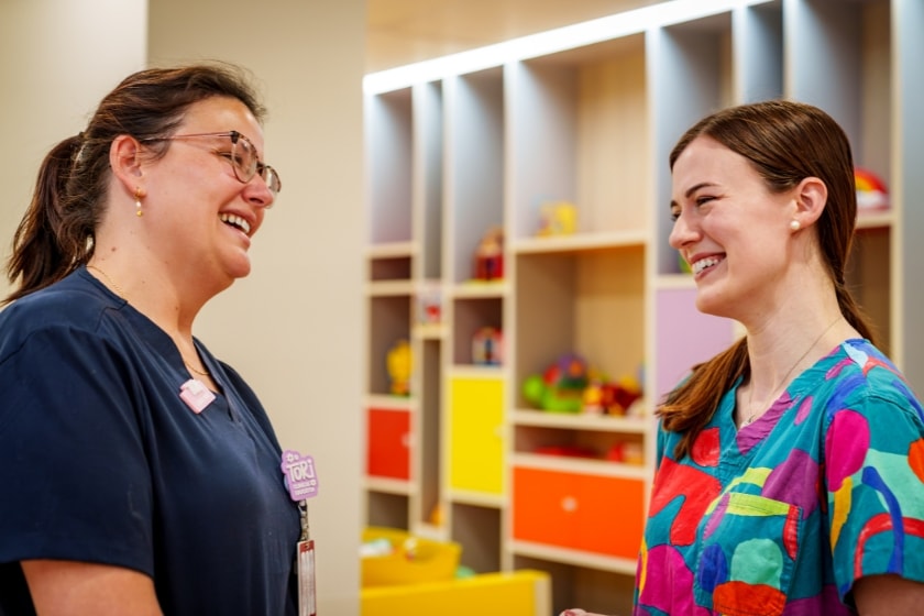 2 nurses are talking to each other with smiles on their face, one of them is wearing a navy blue scrub while the nurse on the right is wearing a colourful scrub.