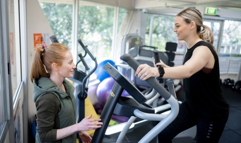 A personal trainer helps train a client on a crosstrainer in a gym.
