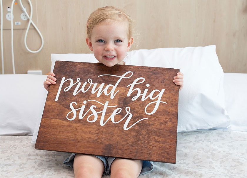A young person sitting on a hospital bed holding a wooden sign saying 