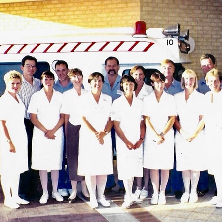 A group of caregivers dressed in uniforms stand in front of an ambulance and the Murdoch Hospital Emergency Department