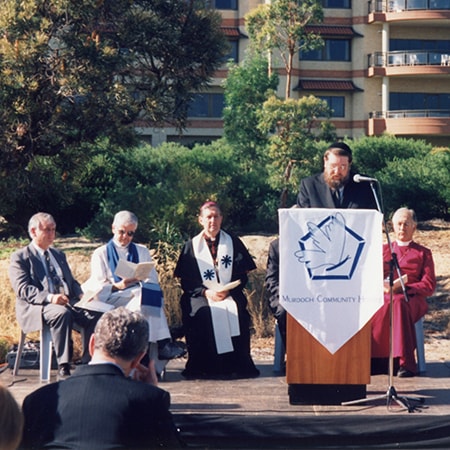 Six religious leaders at a ceremony in front of a crowd in 1997 - Rabbi David Frelich stands at a lectern with a flag draped over it which reads "Murdoch Community Hospice"