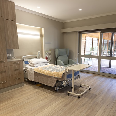 Interior image of a patient room in the Murdoch Community Hospice showing built in cupboards, a bed, a lounge chair and a full length window overlooking a courtyard