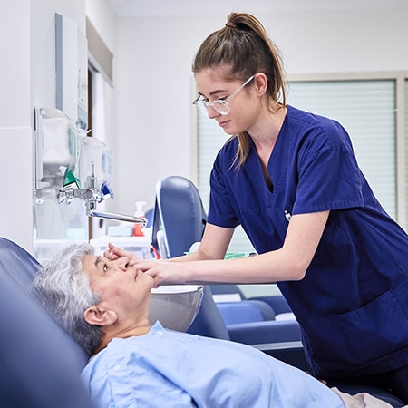 Patient reclined in chair wearing a light blue gown has their eye inspected by a caregiver standing wearing royal blue scrubs