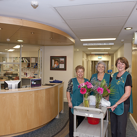 Three volunteers wearing uniform aprons behind a trolley pushing flowers through the St Mary’s Maternity Ward