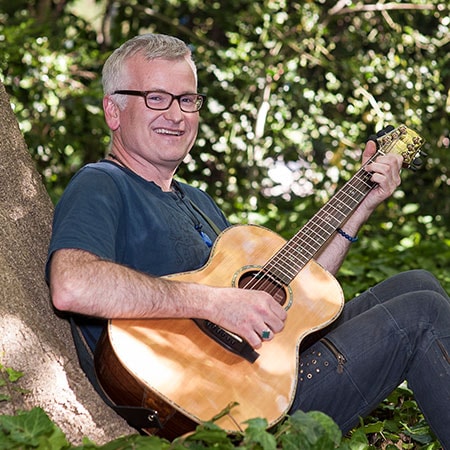NDIS Employment Manager and Positive Behaviour Support Practitioner Chris Milton sitting under a shady tree playing guitar.