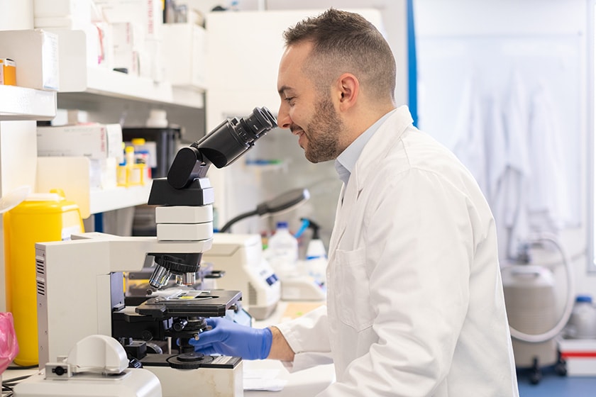 Health professional wearing a coat and disposable gloves in a laboratory looking through a microscope
