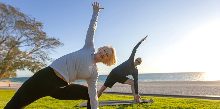 An elderly couple doing a yoga pose next to the beach, stretching their legs wide and reaching one arm to the sky