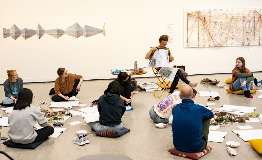 People sitting on the floor looking at the art teacher explain something.