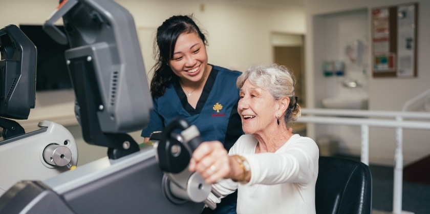 An elderly woman is training on a handcycle with a healthcare professional on her side, both are smiling and enjoying themselves