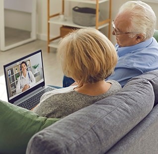 An elderly man and woman view a laptop screen showing a doctor online