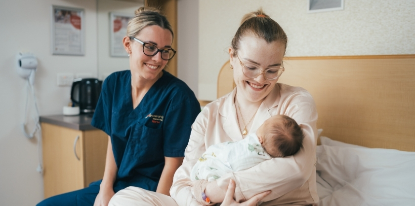 A mother and a midwife lovingly watch a newborn lying in it's mothers arms while sitting on a hospital bed
