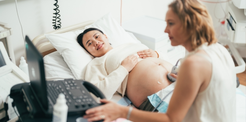 Pregnant woman lying on a hospital bed smiling during an ultrasound examination, while a healthcare professional operates the ultrasound machine beside her.