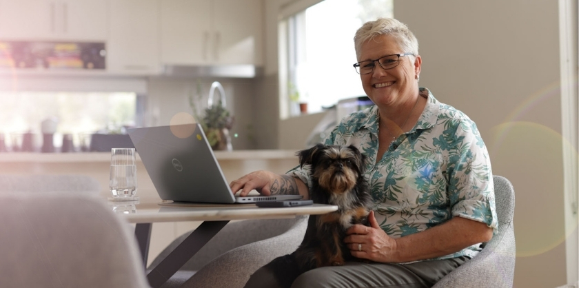 A person sitting behind a computer on a kitchen table with a dog on her lap, smiling at the camera. There are also a glass of water and a phone on the table, in the background there us a blurred, modern kitchen with sun shining through the window