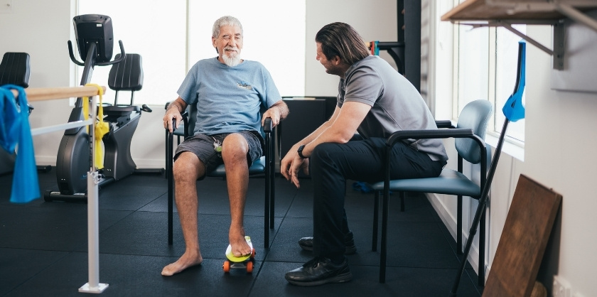 An elderly man is talking to a healthcare professional while having one foot on a skateboard. Both are sitting on chairs in a gym. 