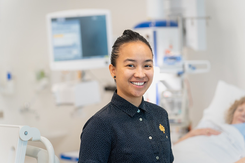 A smiling hospital worker in front of a patient on a hospital bed with some hospital monitors and equipment in the background.