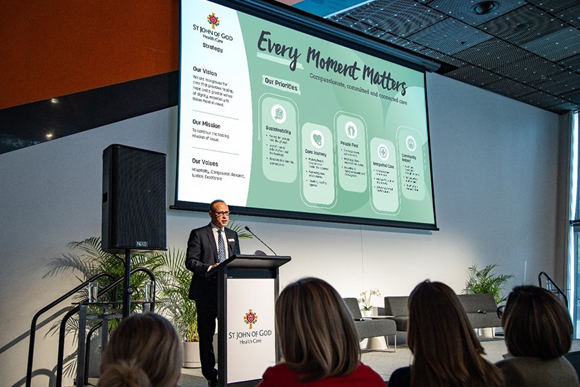 St John of God Health Care CEO Bryan Pyne standing behind a lectern on stage with the St John of God Health Care Strategy's priorities displayed on a screen behind him.