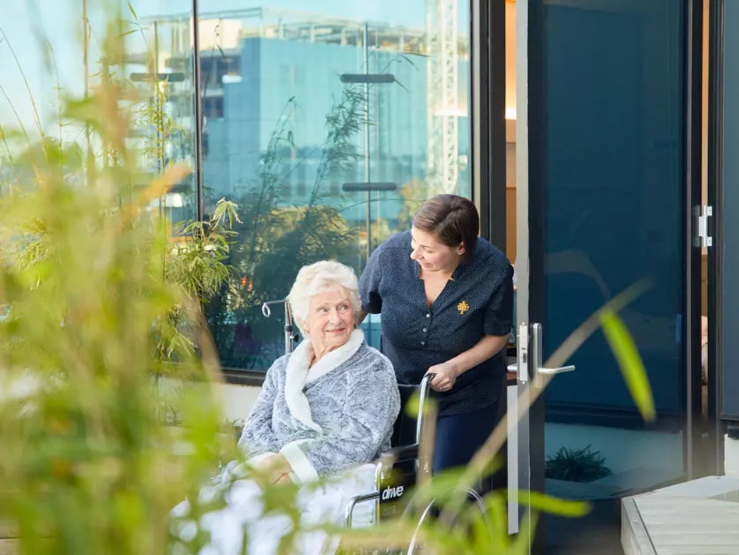 a health professional pushes an elderly lady in a wheelchair out of a building into a lush garden while having a conversation with her