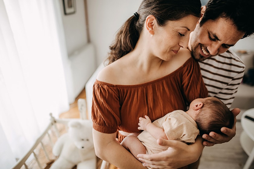 Woman cradling a newborn baby and man leaning over her shoulder cradling the baby's head both glancing down towards the infant smiling whilst standing in a light and airy home nursery.