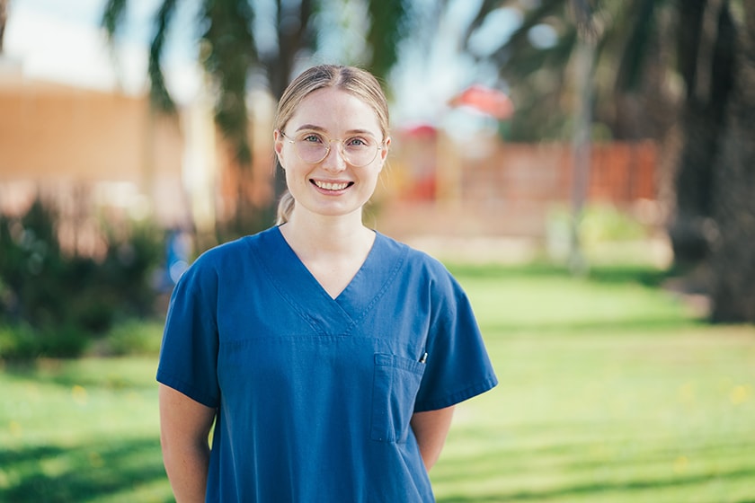 A caregiver wearing blue scrubs standing outside in a park smiling at the camera.