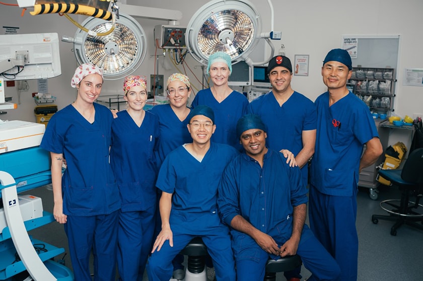 Eight caregivers dressed in blue scrubs in a hospital operating theatre, smiling and looking at the camera.