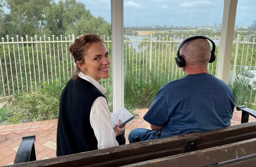 A caregiver smiles at the camera as a patient looks at the view listening to music with a pair of headphones.