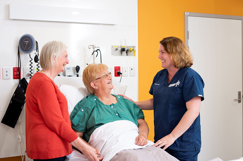 A nurse talks to a patient looking up at her from a hospital bed and while a friend holds the patient's hand