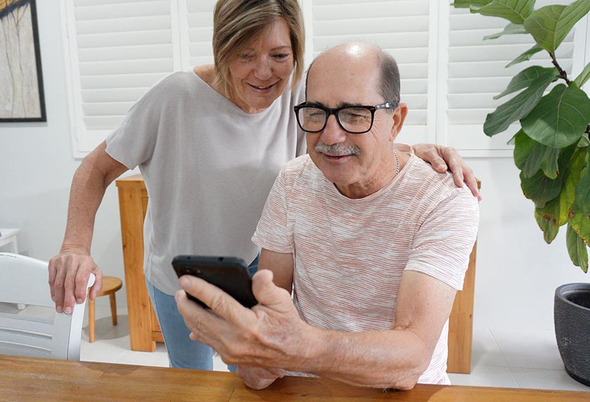 Man sits at table looking at mobile phone screen while a woman stands behind him