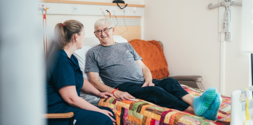 An elderly man lies in an hospital bed while a nurse in a blue scrub sits beside him on a chair, talking to eachother