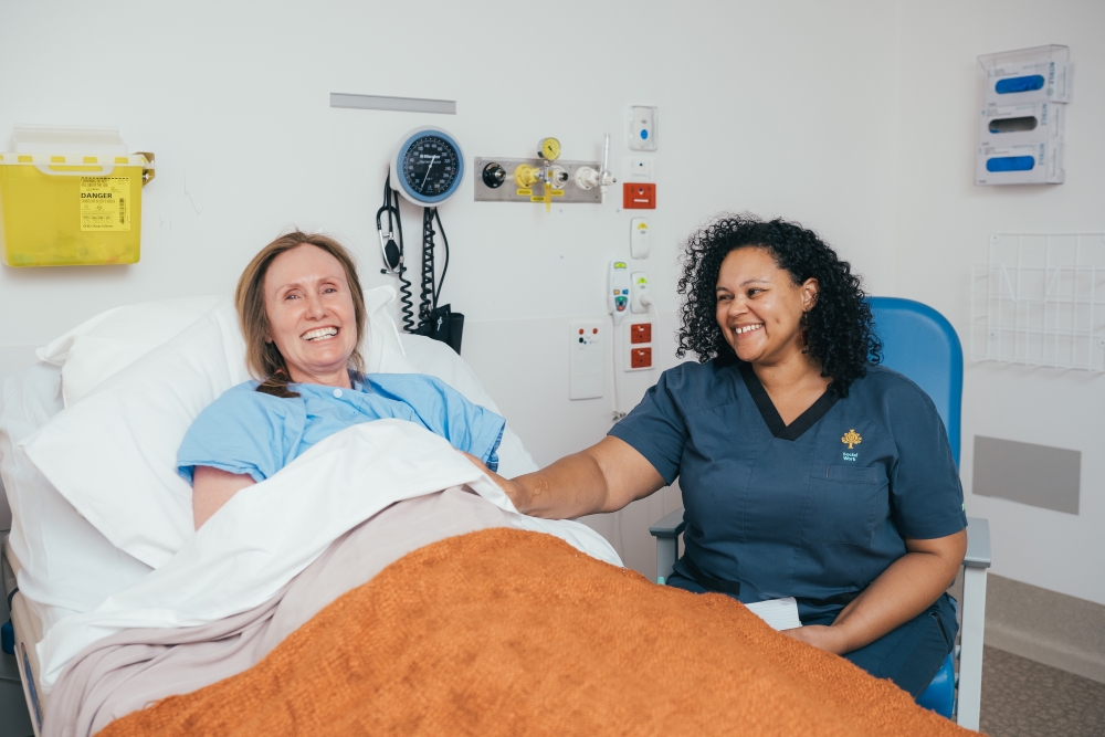 Caregiver sitting next to hospital bed holding patient's hand both smiling