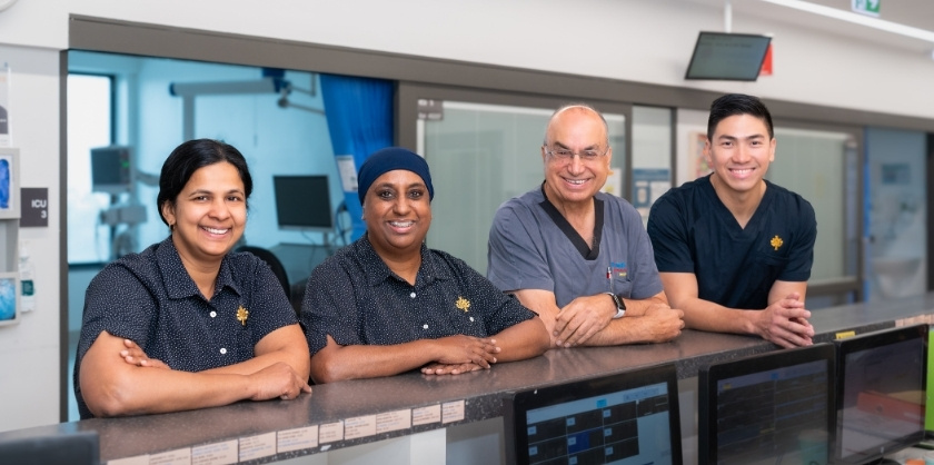 Four healthcare staff members standing together behind a hospital reception desk, smiling at the camera.