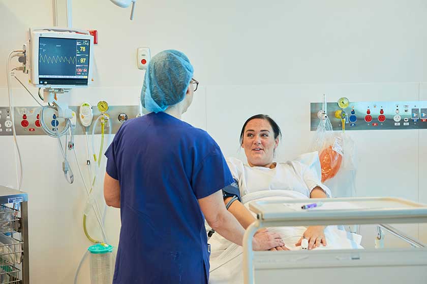 Patient lays in hospital bed wearing gown talking to caregiver in scrubs standing beside them