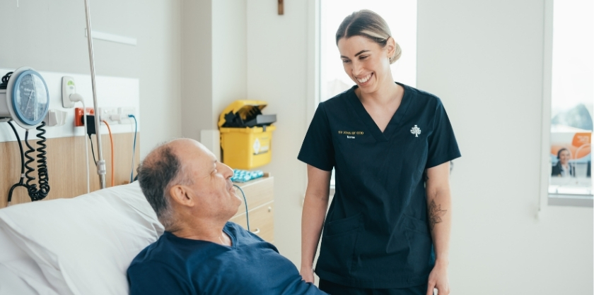 an elderly man lies on a hospital bed with a smiling nurse on his side, both are looking at eachother