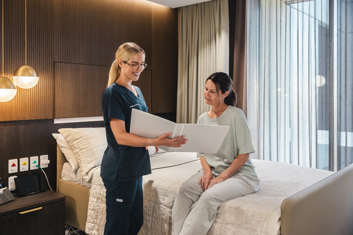 A patient sits on the bed as a caregiver shows her a folder in The Suites.