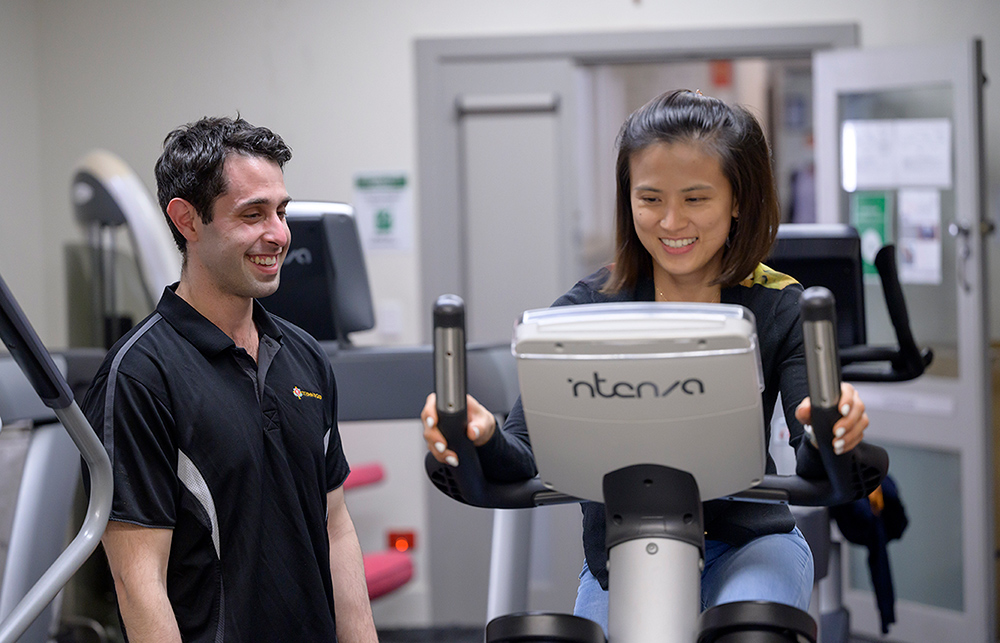 A mental health client uses an exercise bike in the gym with help from a caregiver