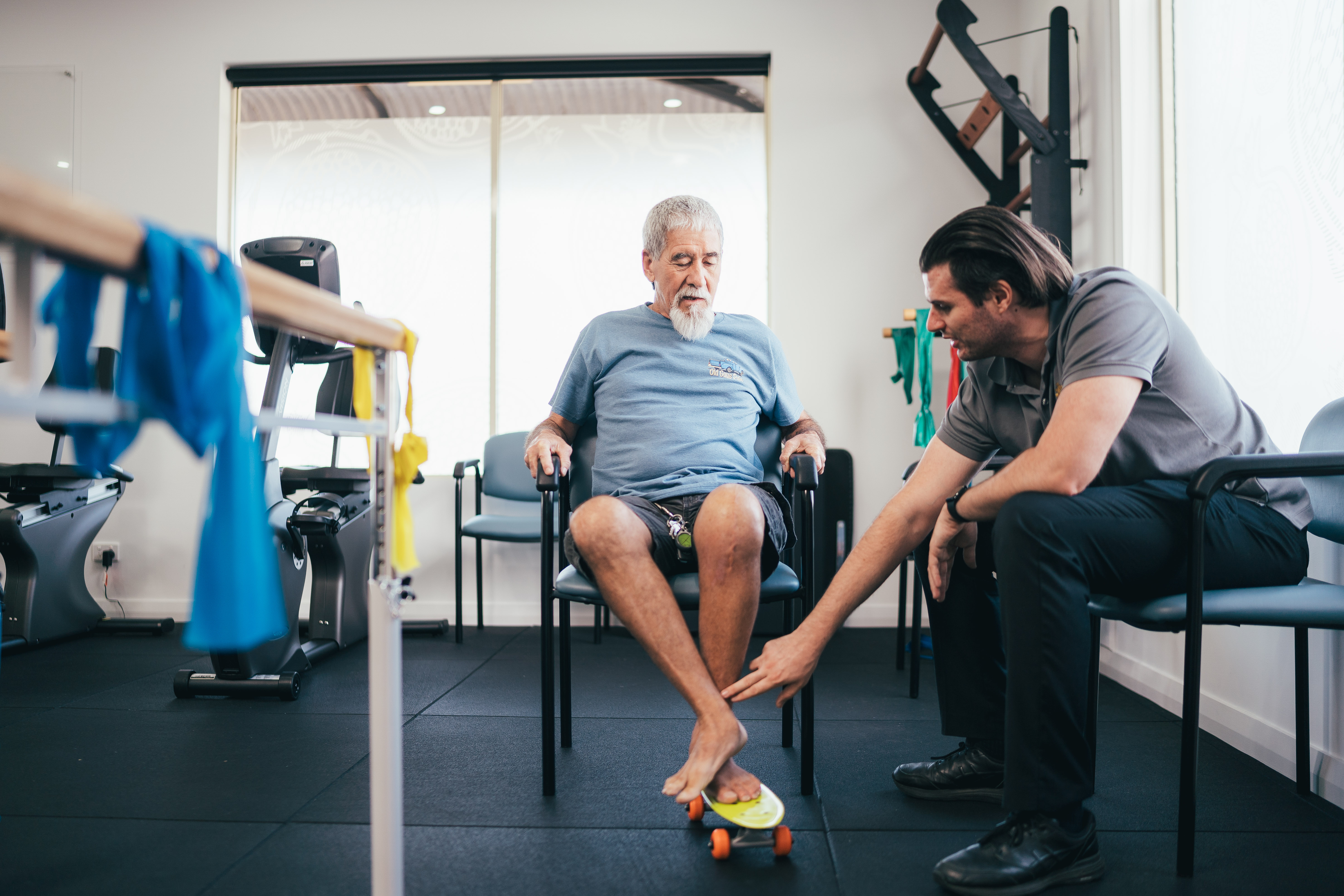 A healthcare worker points to the ankle of a patient that is doing rehabilitation therapy on a skateboard while sitting in a chair