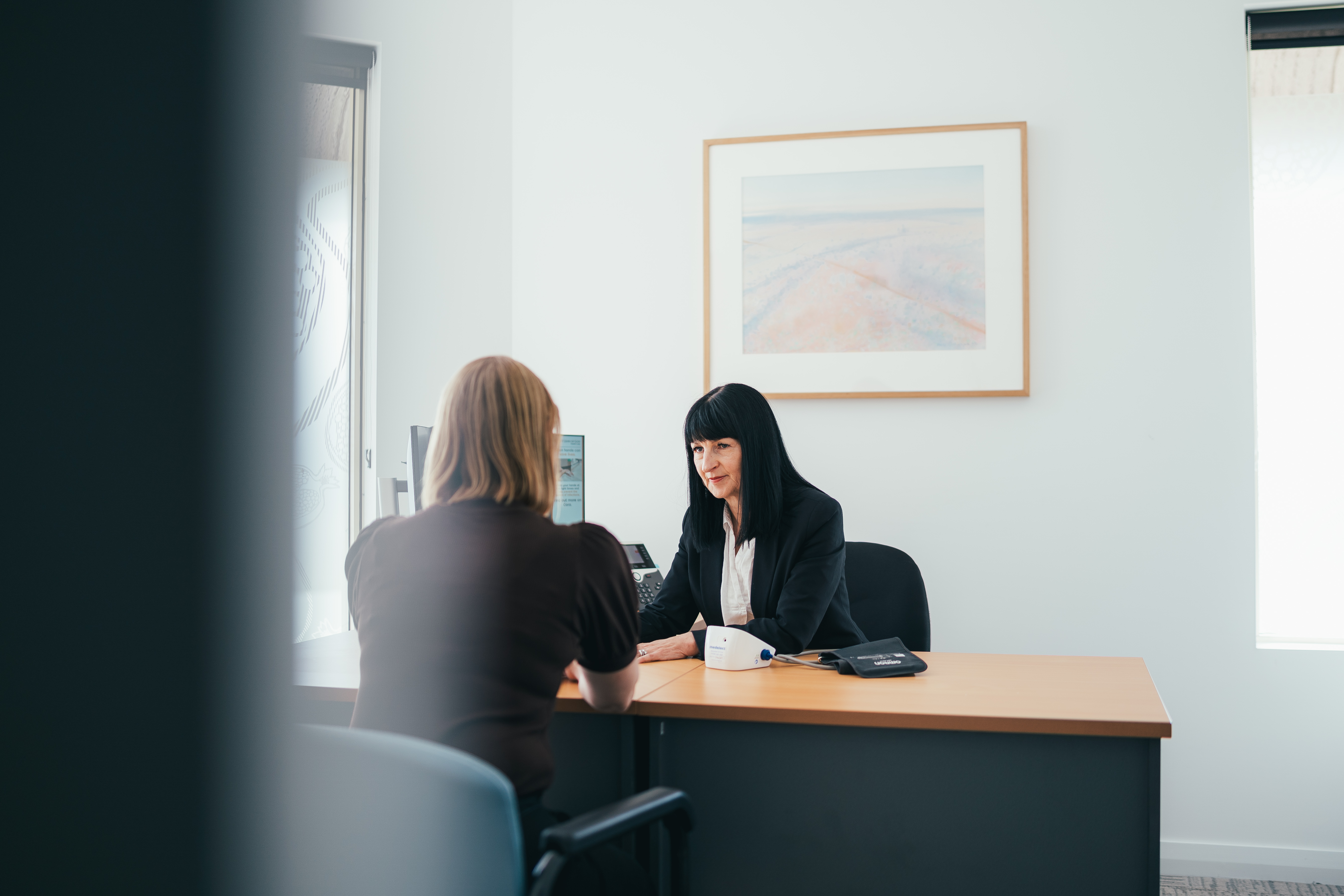 Two people having a conversation over a desk, one person facing the camera and one person with her back to the camera