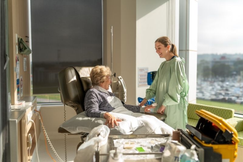 A caregiver chats with a patient sitting in a chair in a hospital room.
