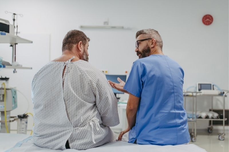 A caregiver has a discussion with a patient sitting on a bed in a hospital room