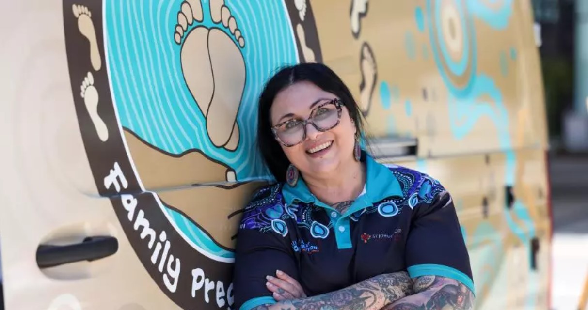 Midwife Jodee who wears a black and blue St John of God polo with aboriginal artwork poses in front of a painted van featuring aboriginal artwork with footprints