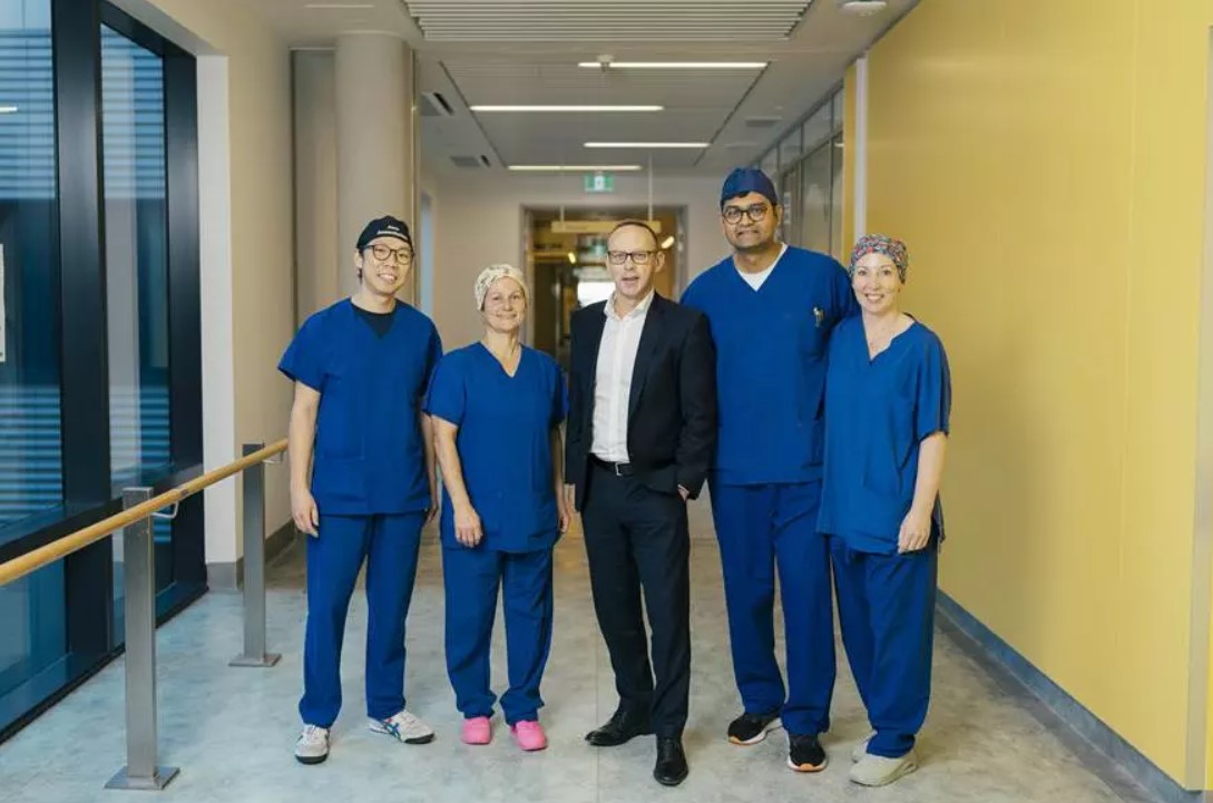 Five people posing in a hospital hallway, the middle person is wearing a suit and the two people flanking him on each side are wearing blue scrubs